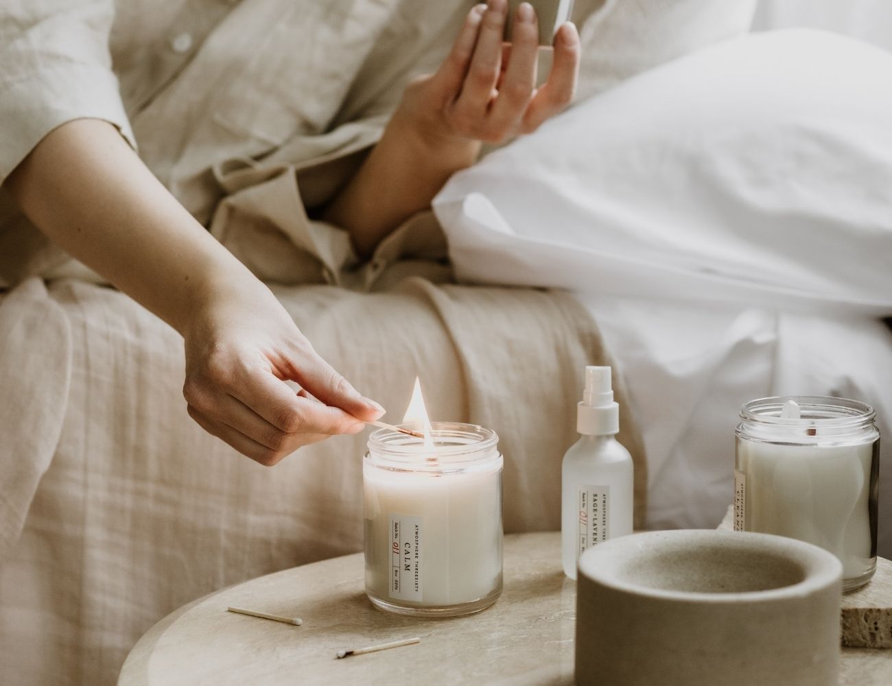 Woman is lighting an Atmosphere Threesixty Aromatherapy Candle on her nightstand, with an Aura Mist sitting next to it.
