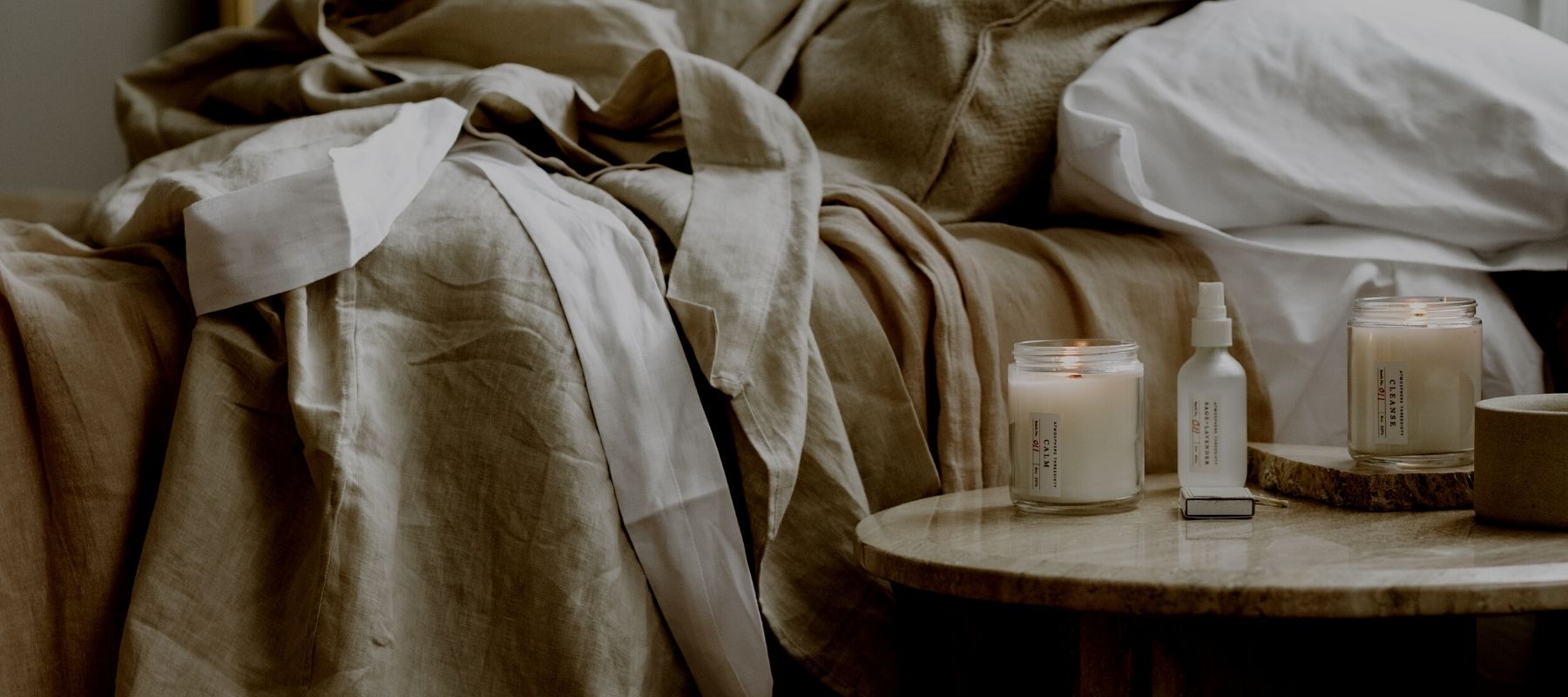 An Atmosphere Threesixty Aromatherapy Candle and Aura Mist are sitting on a bedside table. The bed is unmade.